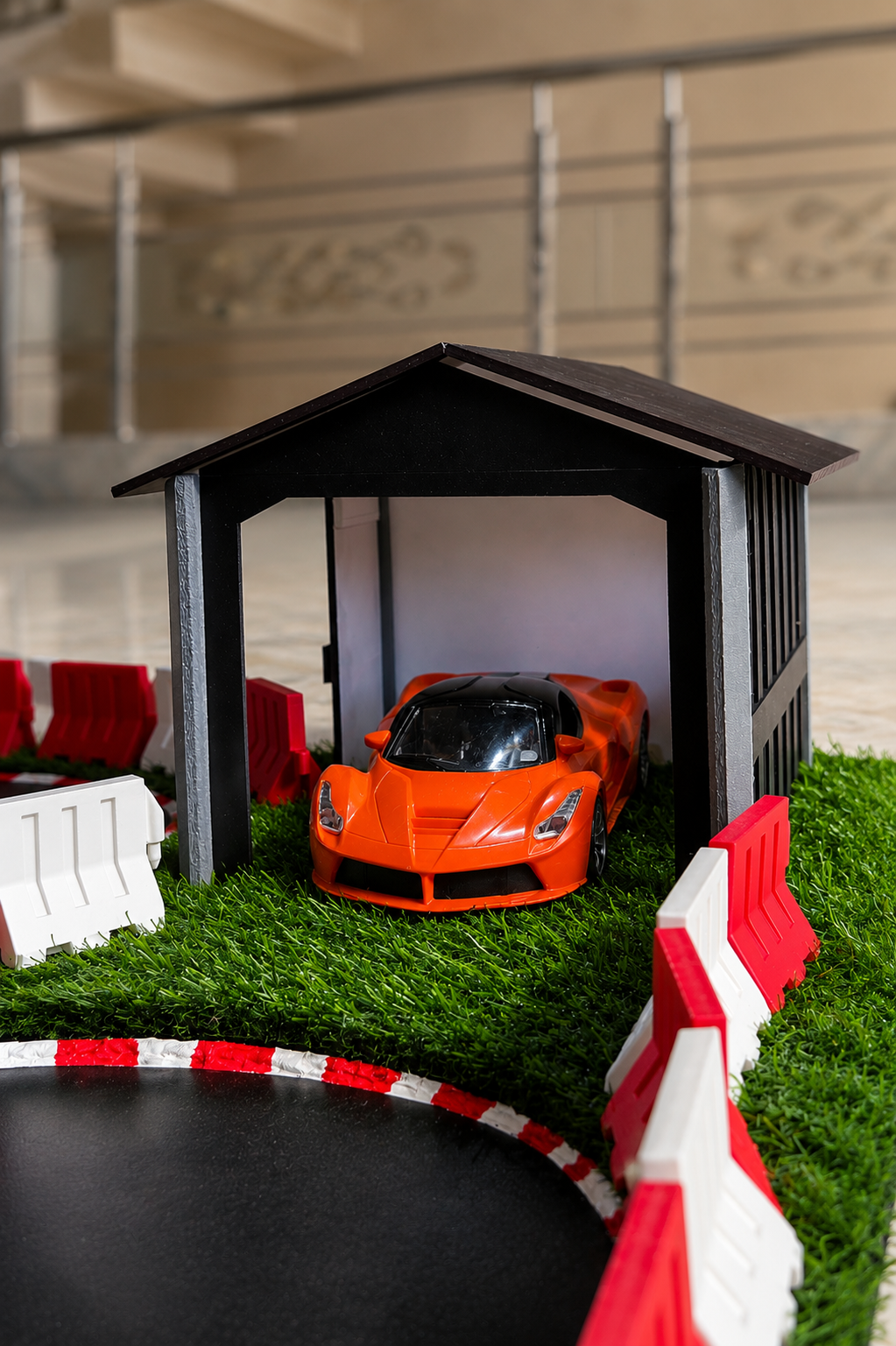 An orange toy sports car parked inside a small black garage structure on artificial green grass, with red-and-white barriers and a blurred indoor background.