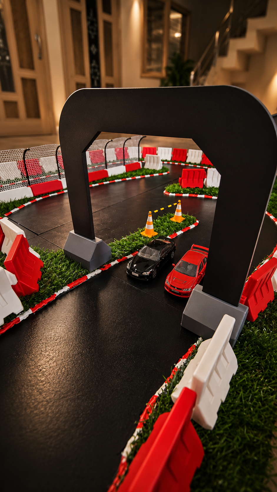 A miniature toy race track setup with a black archway, red and white barriers, artificial grass, and two small cars, photographed indoors in a real room with stairs and windows in the background.