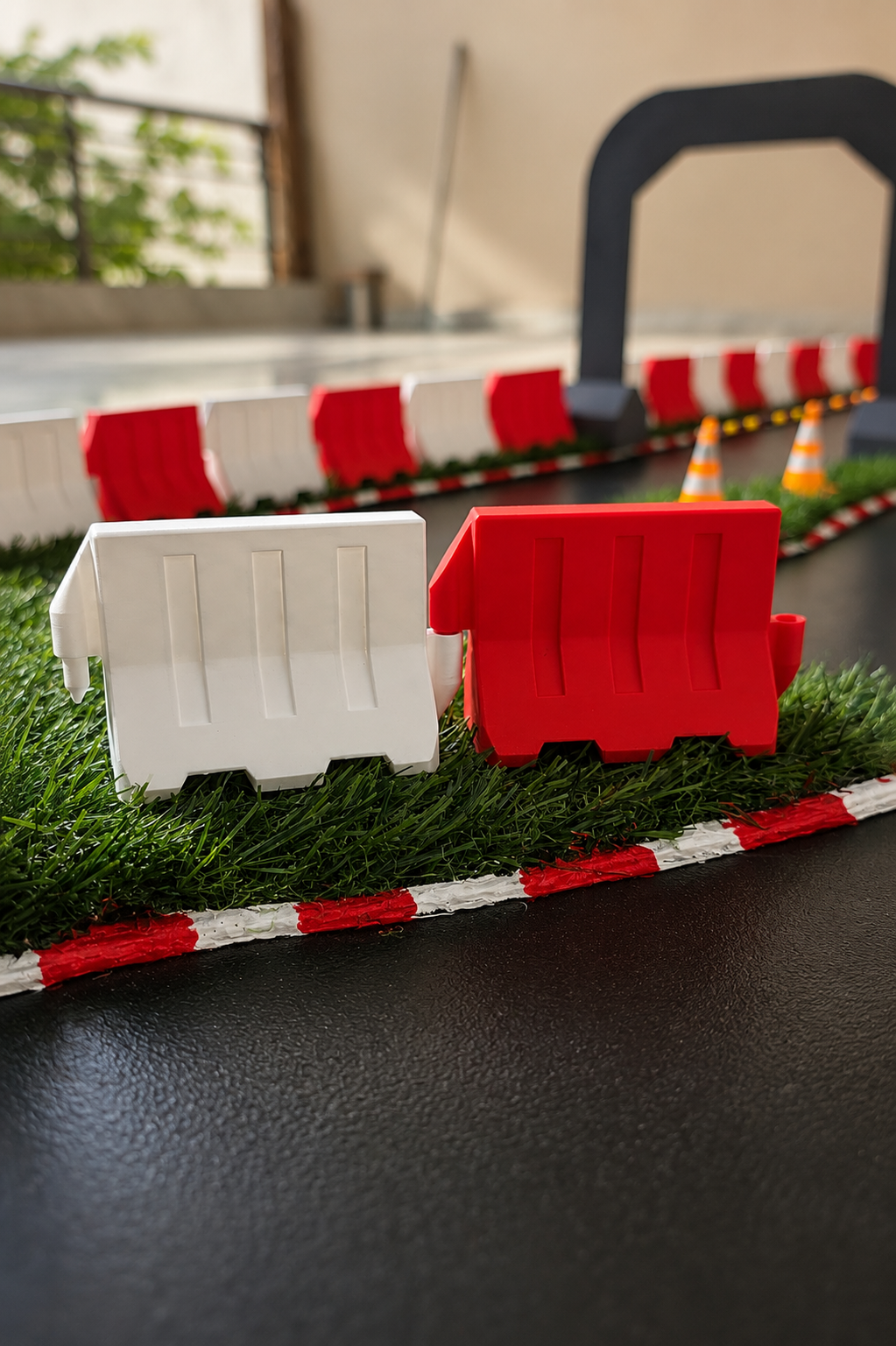 A close-up of a toy construction-themed playset with red and white plastic barriers, artificial grass, and small orange traffic cones on a dark tabletop.