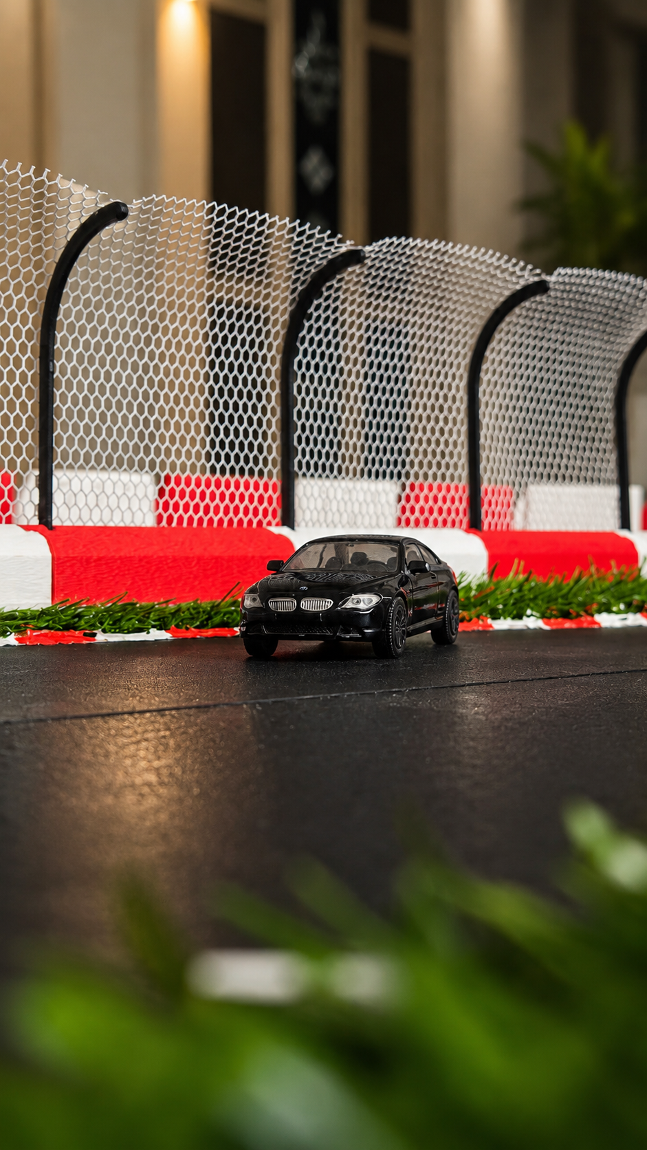 A miniature black toy car model driving on a miniature racetrack with red and white barriers and a safety net in the background.