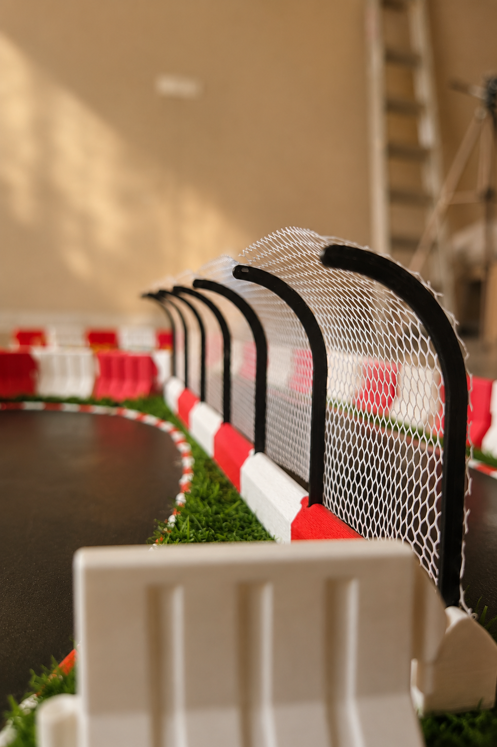 A close-up, angled view of a miniature go-kart track setup, showing a black asphalt-like racing surface, red and white striped barriers, a white safety net, and green artificial grass, with a blurred indoor background.